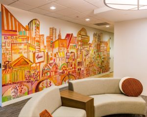 Colorful, hand-drawn Philadelphia cityscape mural spanning an office wall, with abstract buildings in warm orange and pink tones behind curved lounge seating in a modern corporate space.
